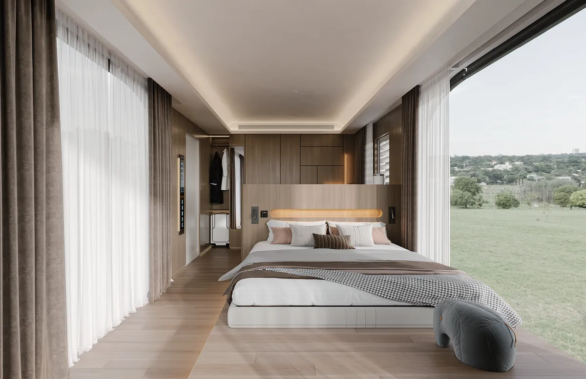 Chambre à coucher moderne avec un grand lit, un parquet en bois et des baies vitrées offrant une vue panoramique sur les champs verdoyants. Des rideaux légers, une chaise grise moelleuse et un éclairage doux créent une atmosphère sereine.